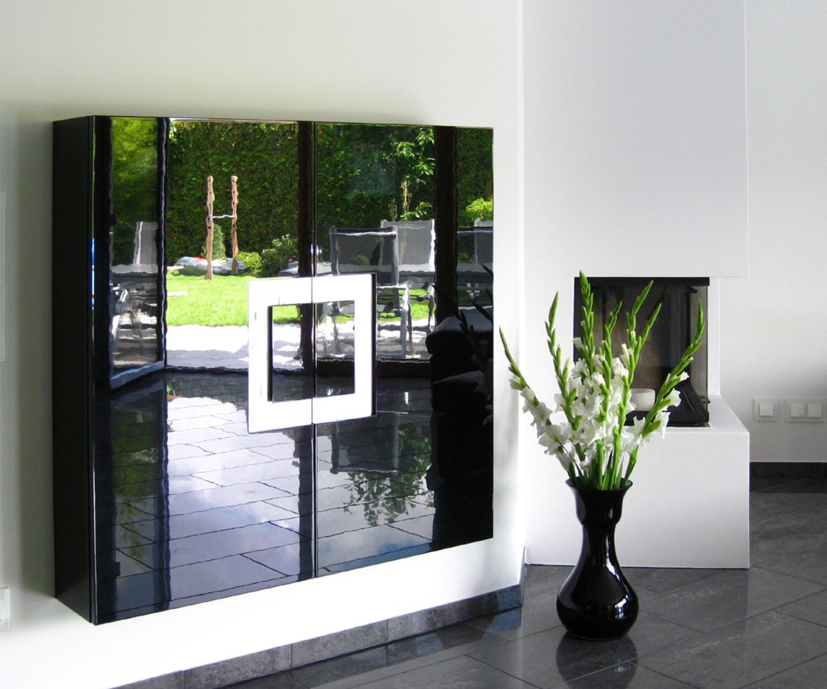 Ein Hängeschrank in geringer Tiefe mit Griffornament in Blattsilber und schwarzer Hochglanzoberfläche. Im Hintergrund ist ein moderner Kamin mit einer Pflanzenvase zu sehen.