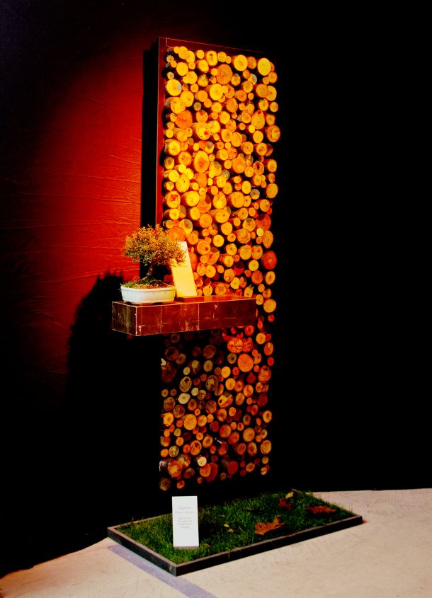 Schmales Wandpaneel mit Rückwand aus Holzscheiben mit Ablage in Blattsilberoberfläche, präsentiert über Kunstrasenstück mit Blattdekoration und dunklem Hintergrund.