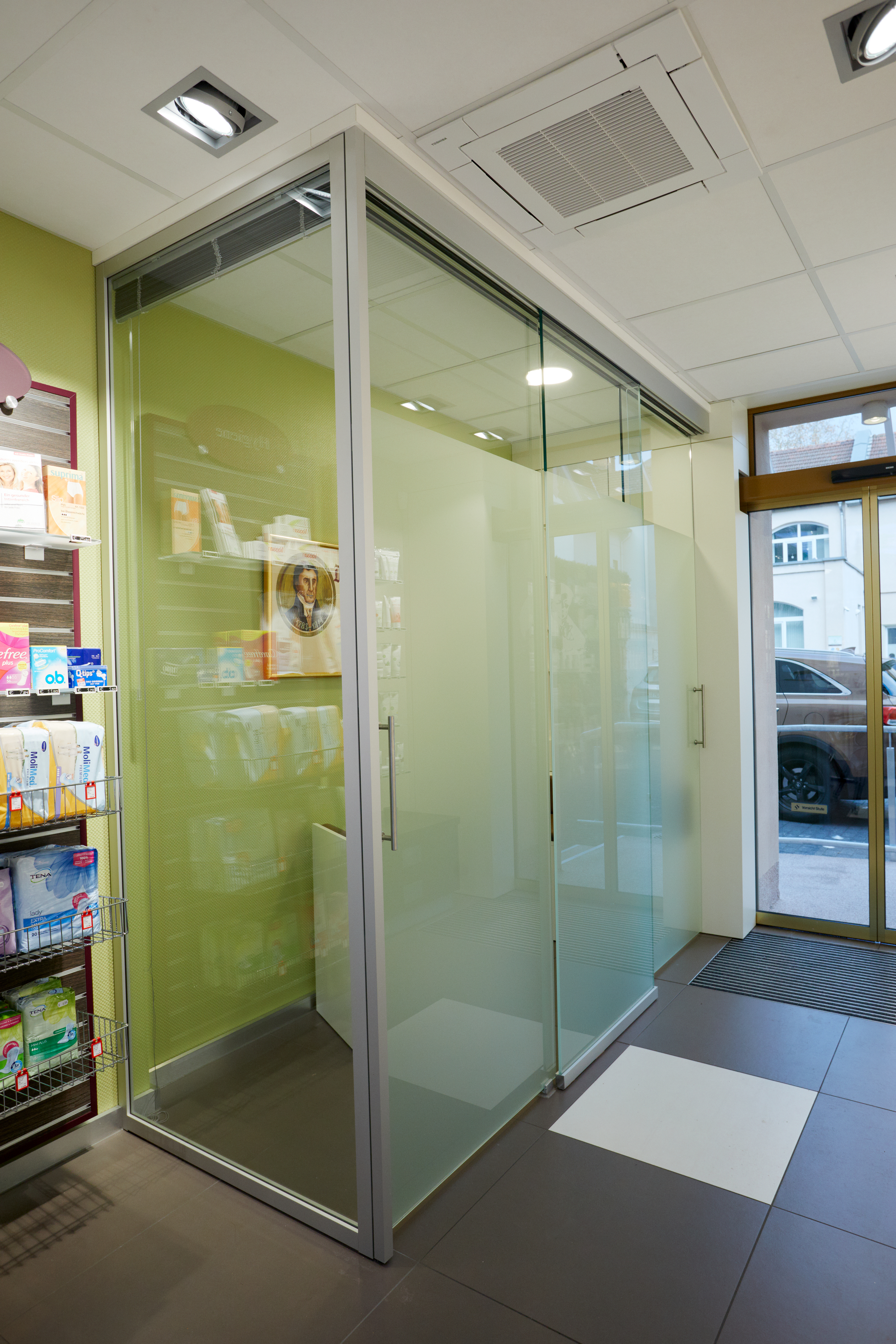 Glasberatungsraum in Apotheke mit Milchglas, grünem Regalbereich, Prospekten, Fliesenboden und Eingangstür im Hintergrund.