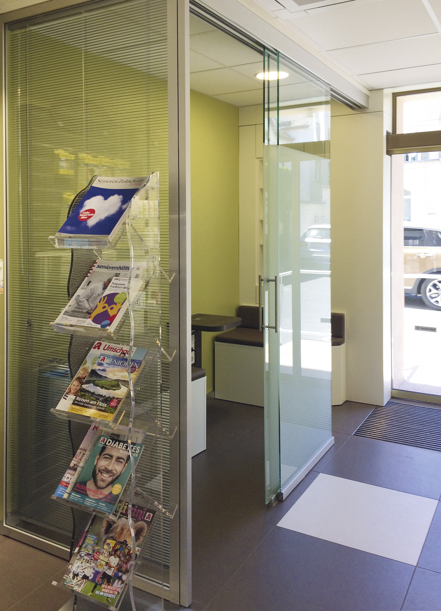 Glasberatungsraum in Apotheke mit Milchglas, grünem Regalbereich, Prospekten, Fliesenboden und offener Eingangstür.