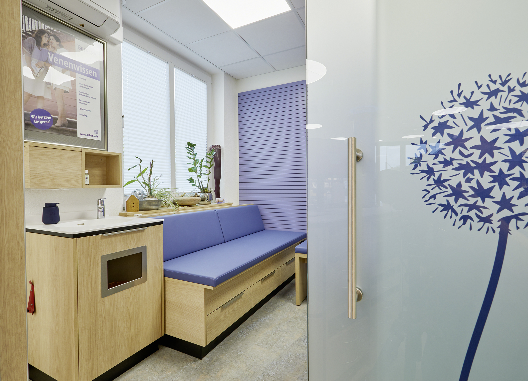 Moderner Beratungsraum mit Holzmöbeln, blauer Sitzbank, Pflanzen, Info-Poster und Glastür mit Pusteblumen-Motiv.