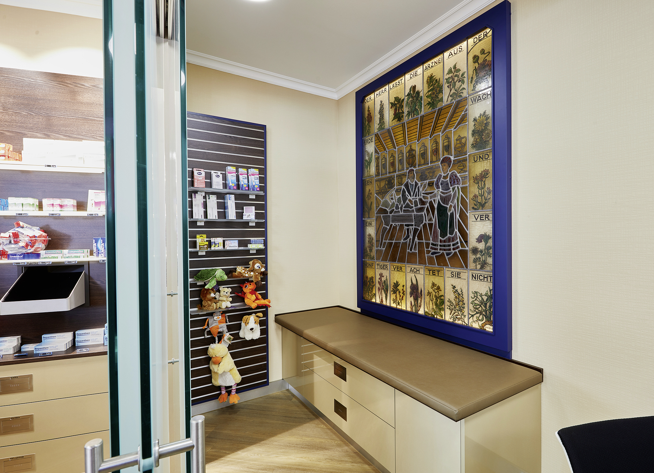 Apotheken-Beratungsraum mit Bank, Regal mit Produkten und Spielzeug sowie beleuchtetem, historischem Kräuter-Glasbild.