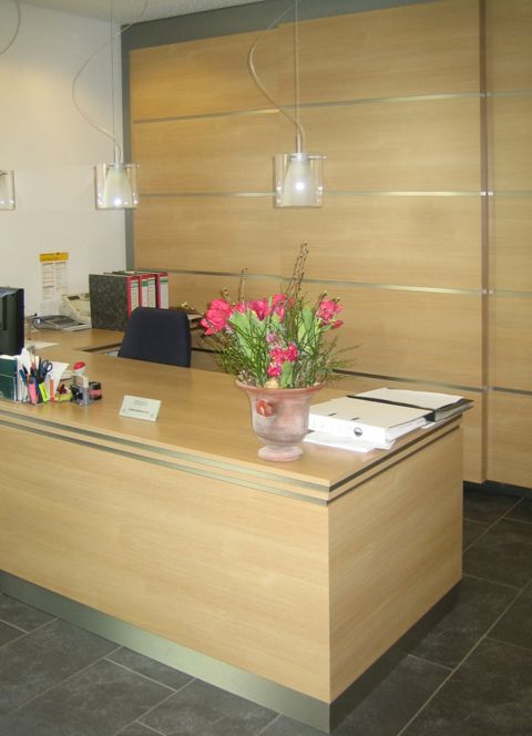 Empfangstheke aus hellem Holz mit Blumenvase, Büroartikeln und Akten, vor passendem Schiebetürenschrank und Pendelleuchten.