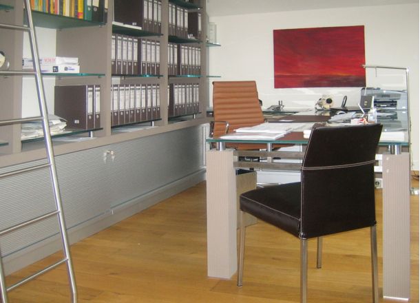 Ein Büroraum mit Schreibtischen aus Glas und verschiedenen Büroutensilien darauf, und großem Wandschrank.