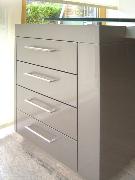 Hochglänzender grauer Büroschrank mit vier Schubladen, Metallgriffen und Glasplatte, auf hellem Steinboden.