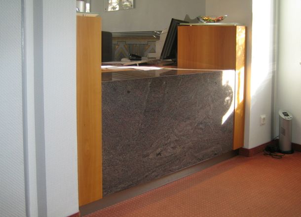 Kompakte Empfangstheke mit dunkler Natursteinfront und seitlichen Holzverkleidungen in einem Büro mit rotem Bodenbelag.