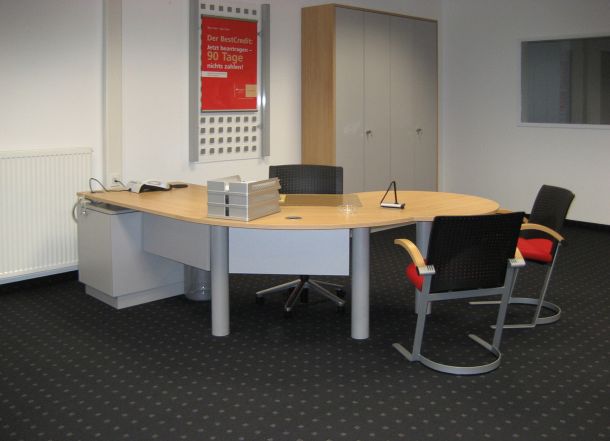 Büroraum mit ovalem Schreibtisch, drei Stühlen, Aktenstapeln, grauen Schränken und rotem Werbeposter an der Wand.