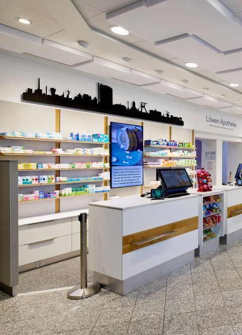 Moderne Apotheken-Offizin mit weißer Verkaufstheke, Warenregalen, digitalem Bildschirm und Oberhausen-Silhouette an der Wand.