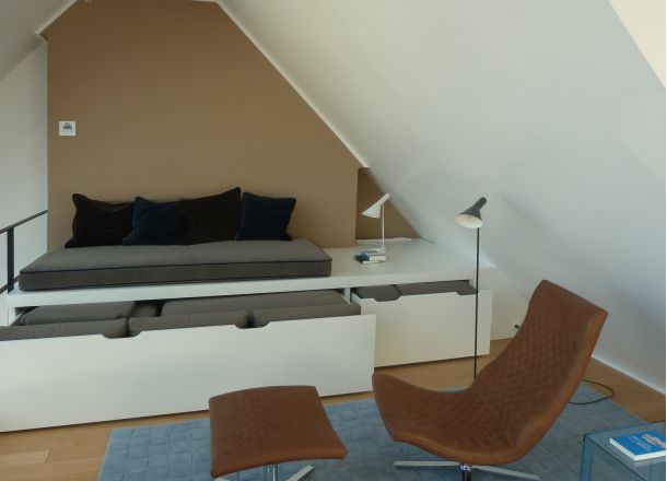 m Vordergund ein Liegesessel mit gepolsterter Fußablage in einem Dachboden-Wohnzimmer, vor einer eingepassten Sitzmöglichkeit mit ausgezogenem Schubkasten für Polster.