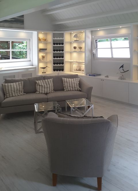 Heller skandinavisch wirkender Wohnraum mit grauem Sofa, Sessel, Glastischen und beleuchtetem Einbauregal mit maritimer Dekoration.