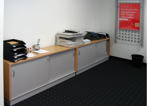 Eine Bürokommode mit Utensilien darauf und geschlossene Schrankfächer, dabein ein Werbeposter.