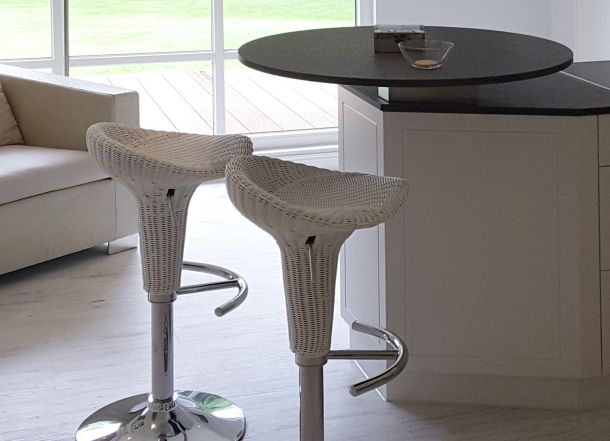 Zwei weiße Barhocker aus Geflecht mit verchromtem Fuß vor rundem Tresen in hellem Wohnbereich mit Sofa und Fensterfront.