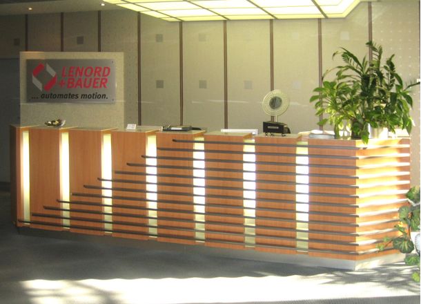 Empfangstheke mit Holzlamellen und Lichtakzenten, Pflanze, Schreibtischzubehör und Logo ‚Lenord + Bauer‘ im Hintergrund.