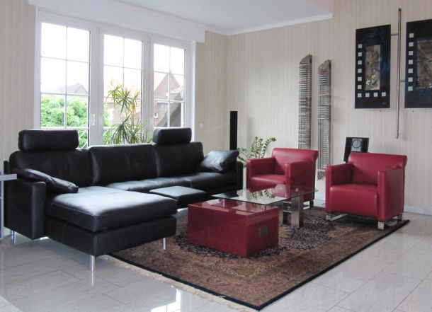 Ein Wohnzimmer mit schwarzer Ledercouch, Sesseln in Weinrot und Glastisch mit Schubladenelement in weinrot gebeiztem, Hochglanz lackierten Vogelaugenahorn.