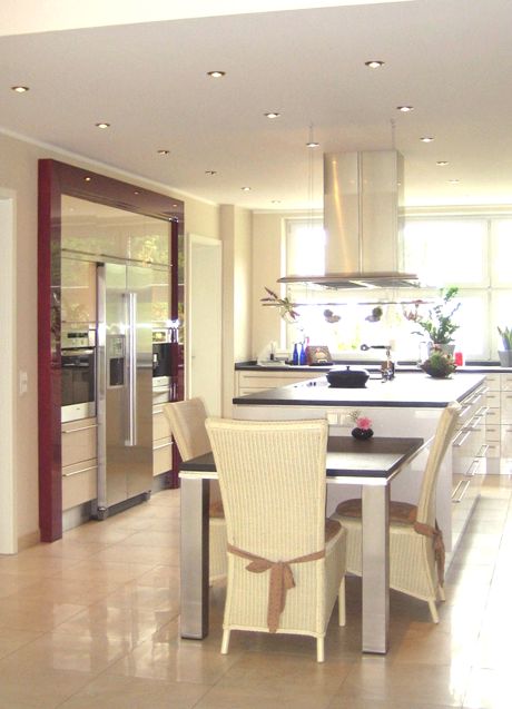 Moderne cremefarbene hochglänzende Küche mit Kochinsel, Dunstabzug, Esstisch, vier Korbstühlen, Edelstahlgeräten und großen Fenstern. Die Gerätewand wir von einem dunkelrot gebeizten, hochglänzendem Ahornrahmen eingefasst.