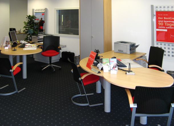 Moderner Büroraum mit mehreren Schreibtischen, schwarzen Stühlen mit roten Polstern, Technik, Pflanzen und Werbepostern.