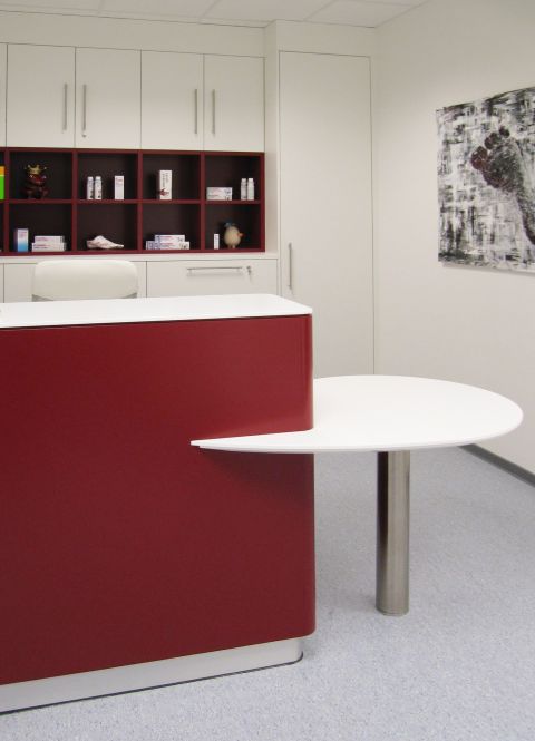 Ein moderner Empfangsbereich mit Theke und integriertem, runden Beratungstisch. in weiß und weinrot. Im Hintergrund eine Schrankwand in weiß mit offenem Regalbereich in weinrot.