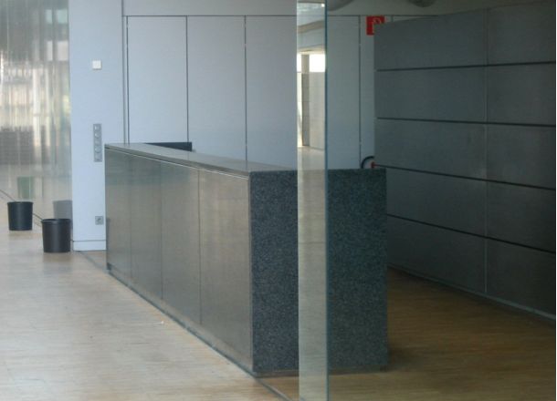 Minimalistische Empfangstheke aus dunklem Stein in modernem Büroflur mit Holzboden, Glaselementen und grauen Wandpaneelen.