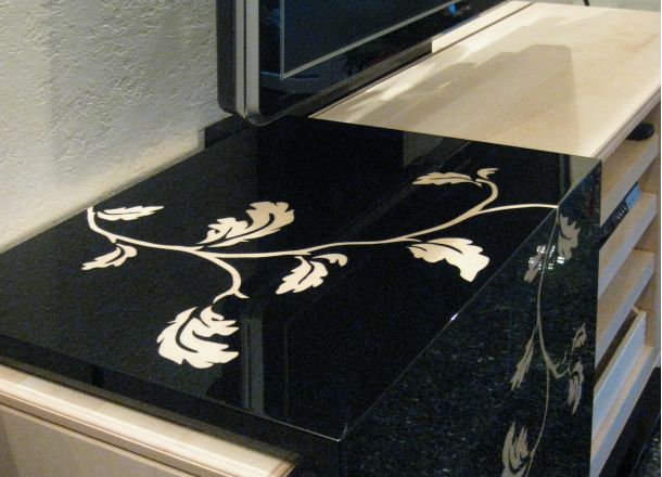 Schwarzes Hochglanzmöbel mit floraler Intarsie in heller Holzoptik, angrenzend an TV-Bereich und helle Schubladenelemente.