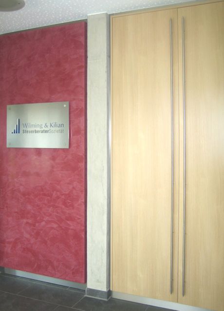 Eingangsbereich mit roter Strukturwand, Kanzleischild ‚Wilming & Kilian SteuerberaterSozietät‘ und hoher Holztür mit Griffleisten.