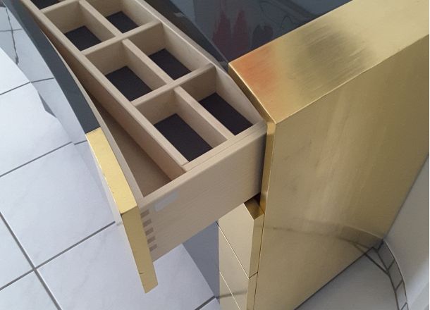 Detailaufnahme eines goldfarbenen Schranks mit geöffneter Schublade und edler Inneneinteilung auf glänzendem Fliesenboden.