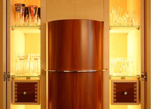 Beleuchtetes Vitrinenmöbel mit Gläsern, zwei Schubladen und zentralem Element aus hochglänzendem Holz mit Metallband.