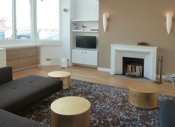 Wohnzimmer mit Einbau TV-Möbel, Heizkörper Verkleidungen mit Eicheholzlamellen, goldenen Tisch-Hockern, Kamin, schwarzer Couch und TV während die Fenster im Hintergrund einen Ausblick auf die Dächer von Paris gewähren.