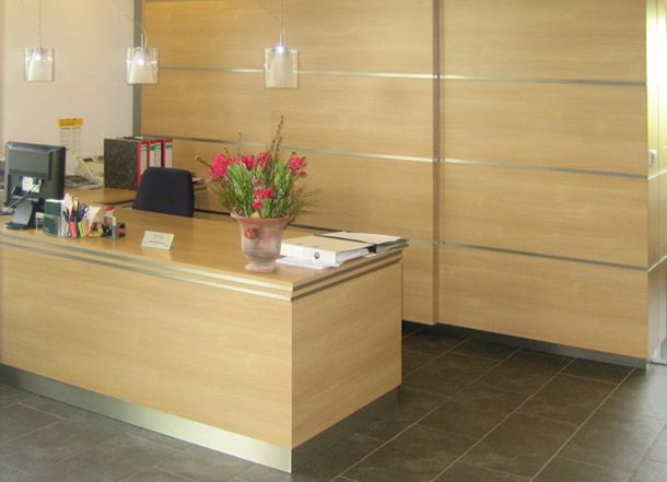 Empfangsbereich mit hellholzfarbener Theke, Blumentopf, Akten, Pendelleuchten und passender Wandverkleidung auf Fliesenboden.