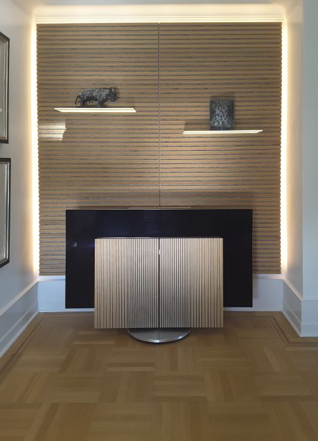 B & O Fernseher Beovision Harmony mit vertikalen Holzlamellen vor beleuchteter Lamellen Holzrückwand horizontal mit zwei schwebenden Regalböden und Dekoobjekten.