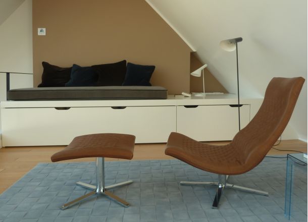 Im Vordergund ein Liegesessel mit gepolsterter Fußablage in einem Dachboden-Wohnzimmer, vor einer eingepassten Sitzmöglichkeit mit Stauraum für Polster.