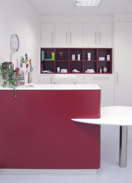 Empfangstheke in Rot mit weißer Auflage, dahinter Hochschränke und offene Regale mit Produkten in heller Praxisumgebung.