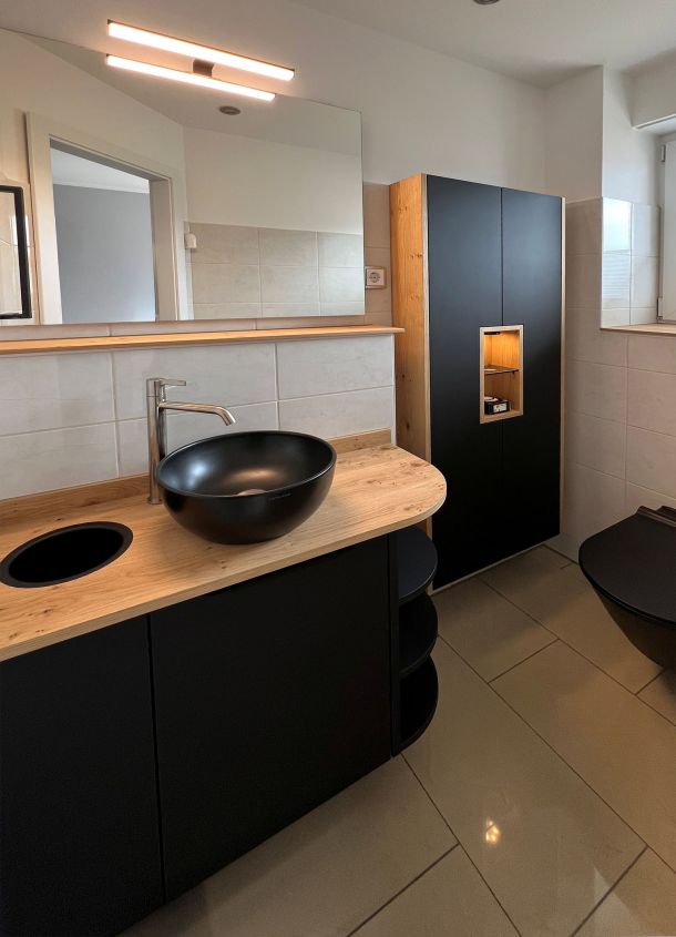 Badezimmer mit Waschtisch in schwarz mit Aufsatzschale in schwarz und Handtucheinwurf, Deckblatt in Massivholz. Im Hintergrund ein Hängeschrank in schwarz mit Massivholzseiten und beleuchtetem Massivholzfach.